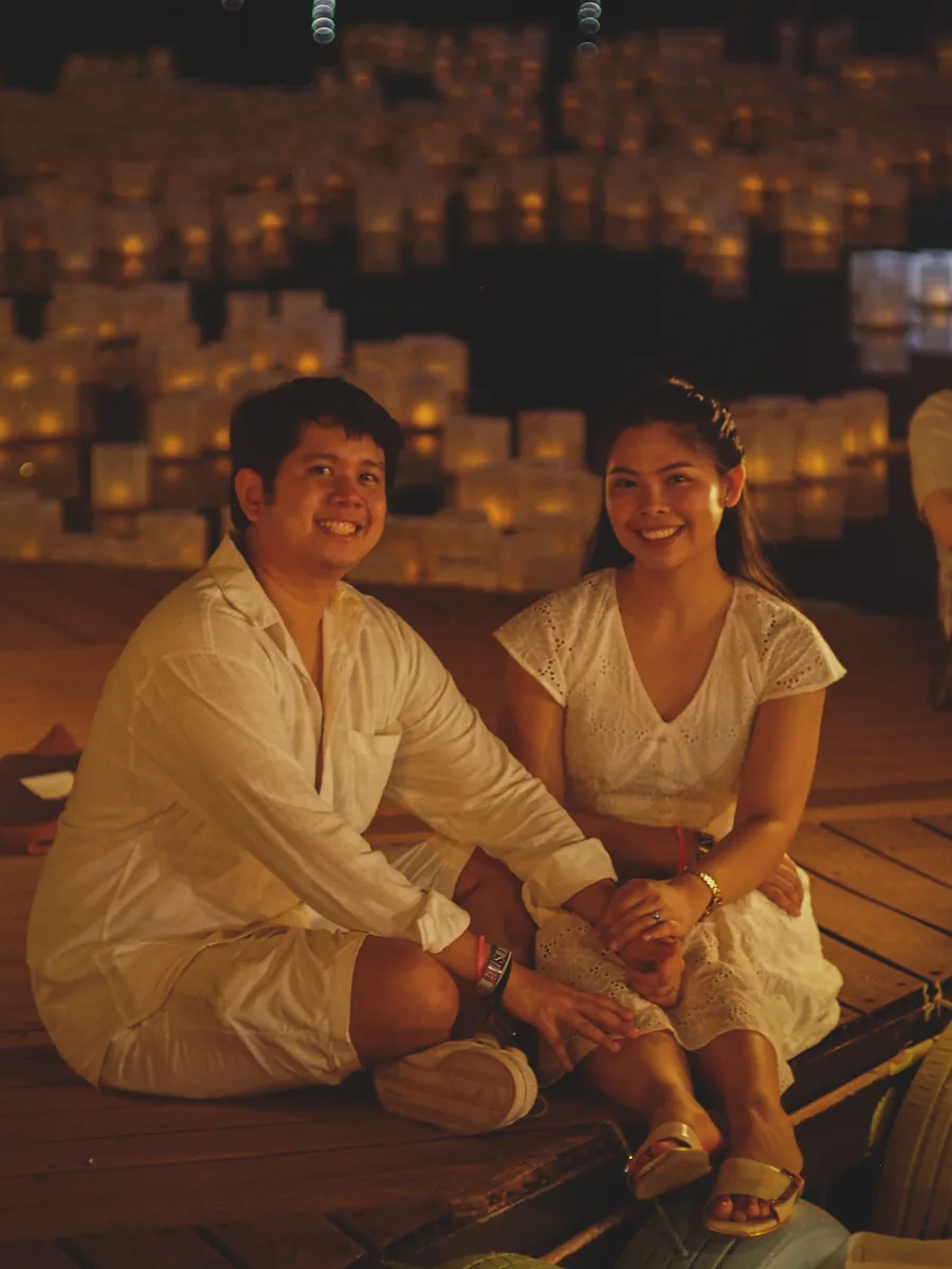 Tommy and Jeyan sitting together on a lakeside dock surrounded by floating lanterns